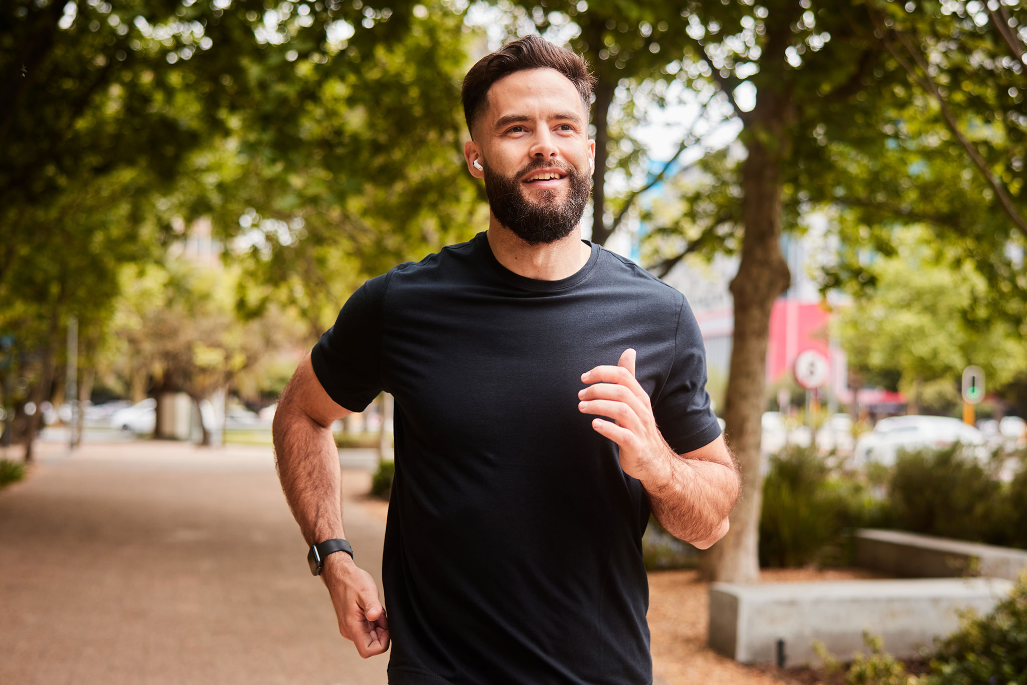 Smiling young man in sportswear listening to music on wireless earbuds while running along a tree lined footpath in an urban park